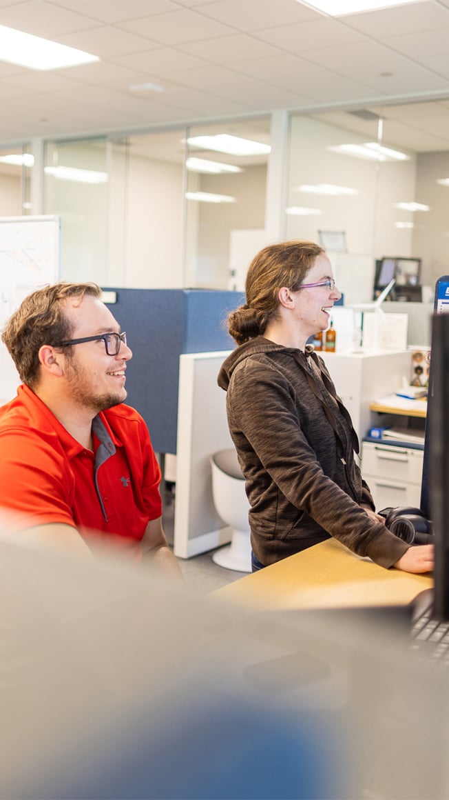 male and female Caucasian engineers working in a corporate office building, looking at computers and smiling